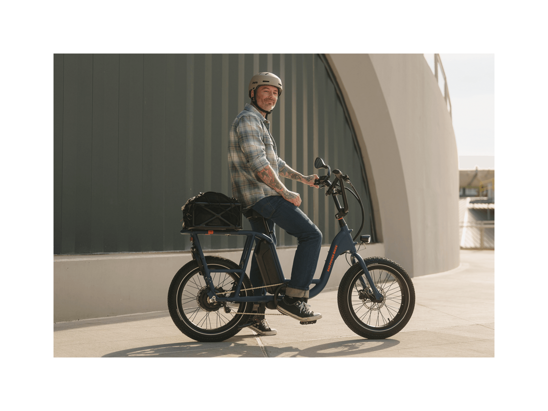 A man with tattoos wearing a plaid shirt, jeans, and a grey helmet smiling and sitting on an ebike in an urban setting. 