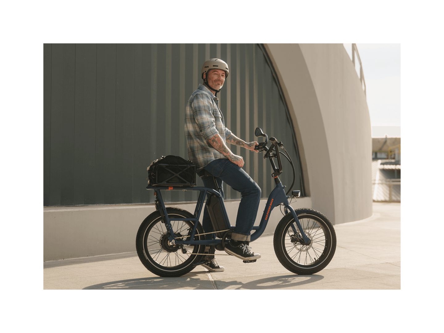 A man with tattoos wearing a plaid shirt, jeans, and a grey helmet smiling and sitting on an ebike in an urban setting. 