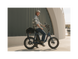 A man with tattoos wearing a plaid shirt, jeans, and a grey helmet smiling and sitting on an ebike in an urban setting. 