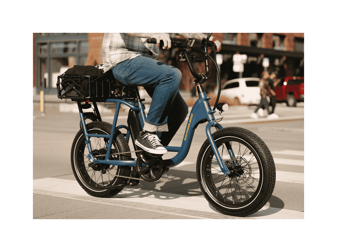 A man riding a blue electric bicycle with an orange Rad Power Bikes logo, fat black tires, and large basket with a cargo net, on a paved road in an urban setting. 