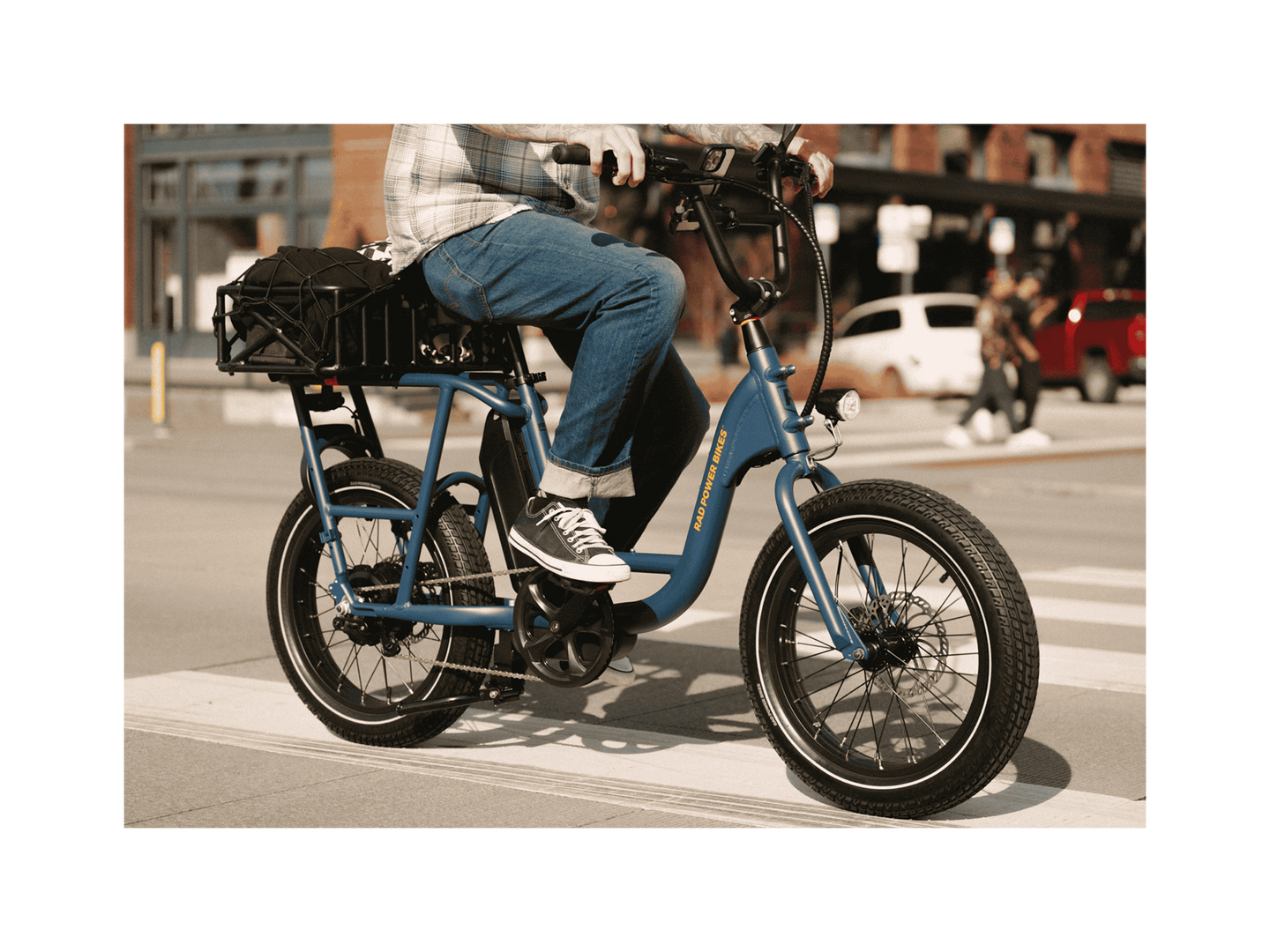 A man riding a blue electric bicycle with an orange Rad Power Bikes logo, fat black tires, and large basket with a cargo net, on a paved road in an urban setting. 