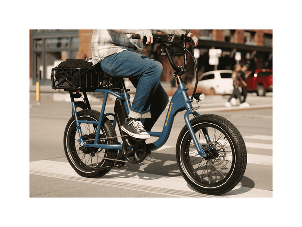 A man riding a blue electric bicycle with an orange Rad Power Bikes logo, fat black tires, and large basket with a cargo net, on a paved road in an urban setting. 