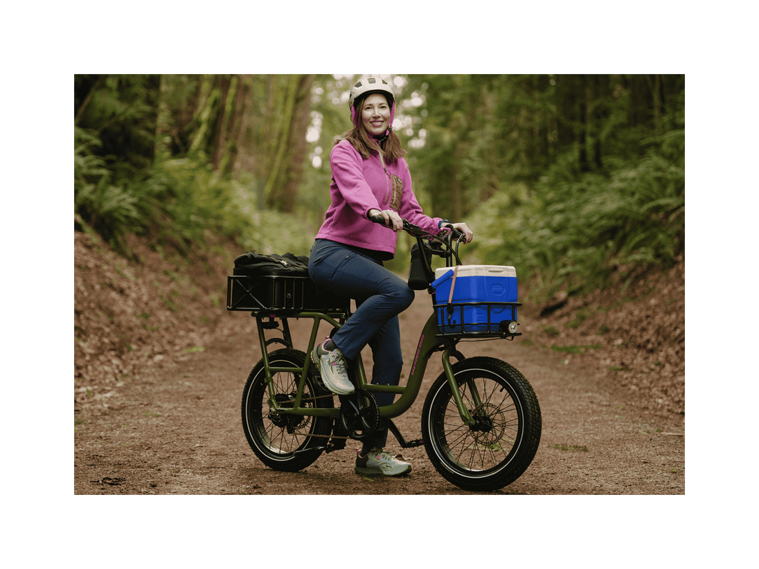 A woman on a trail in a forest sitting on a green cargo ebike with a small front basket and large rear basket wearing a pink jacket and white helmet.
