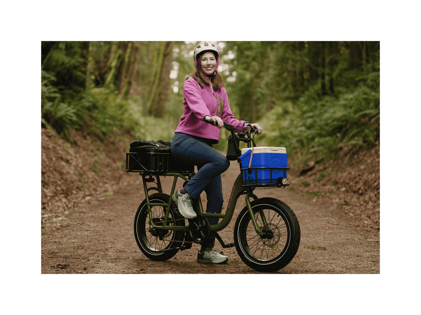 A woman on a trail in a forest sitting on a green cargo ebike with a small front basket and large rear basket wearing a pink jacket and white helmet.