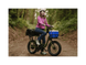 A woman on a trail in a forest sitting on a green cargo ebike with a small front basket and large rear basket wearing a pink jacket and white helmet.